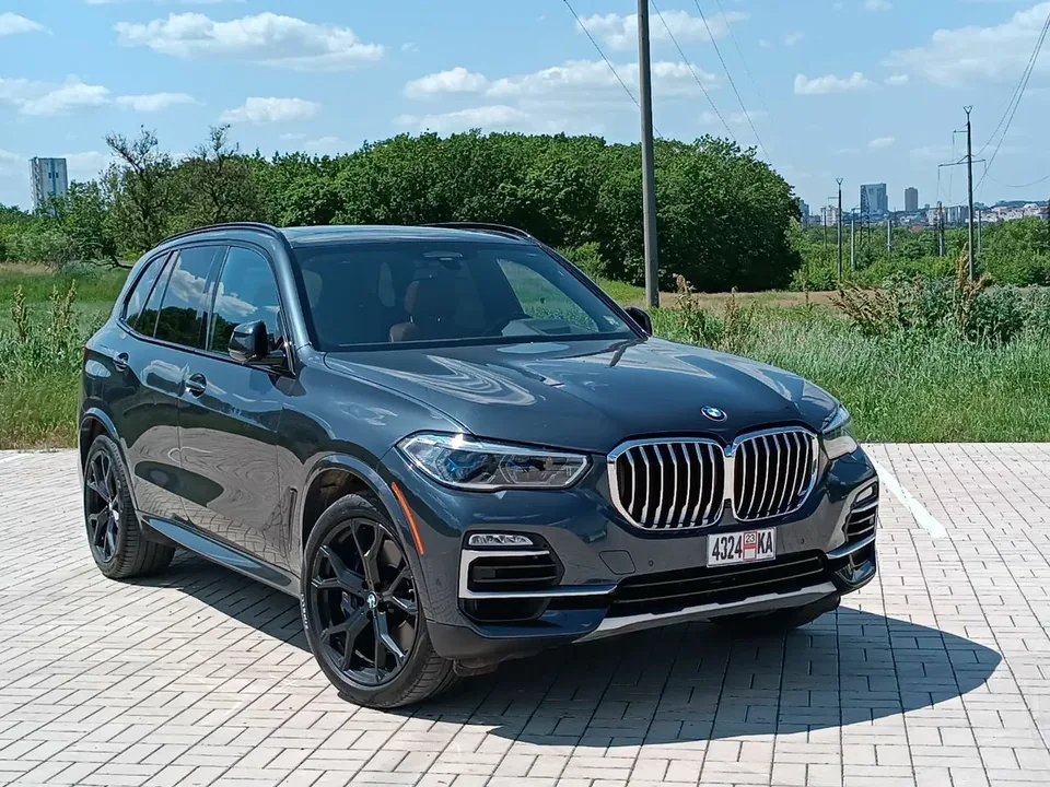A dark grey BMW X5 on a paved lot, trees behind it
